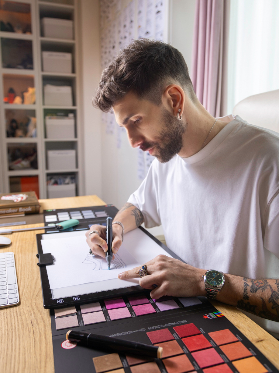Luca Sartori al lavoro su uno schizzo di una scarpa in uno studio di design, con una tavolozza di colori e bozzetti appesi sullo sfondo.